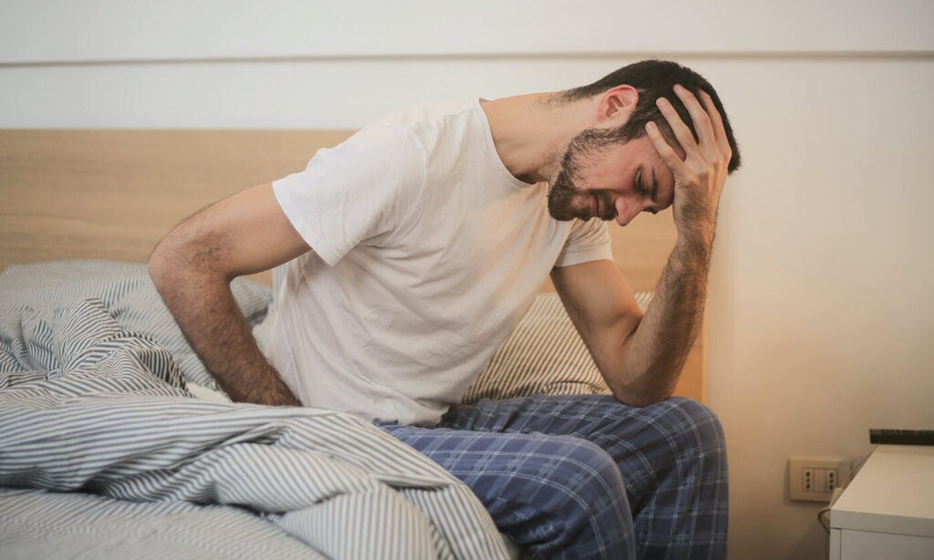 hangover Person hungover sitting on the edge of a bed holding his head